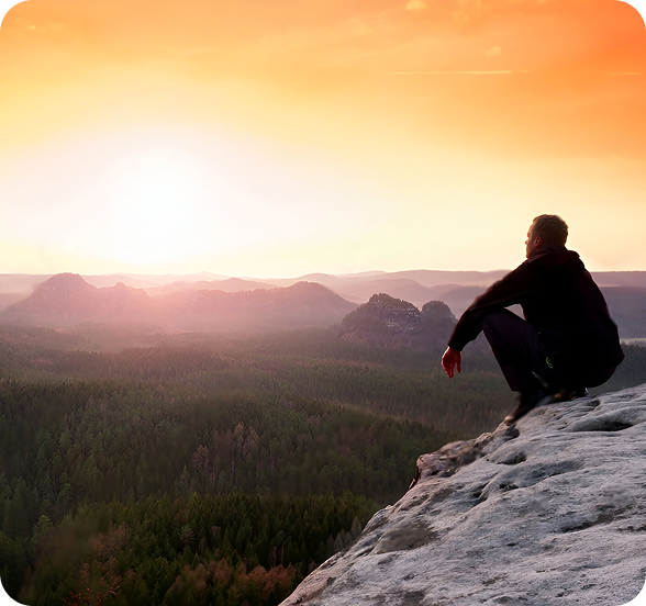 A man sits upon the peak of a mountain staring into the distance at the sun rising