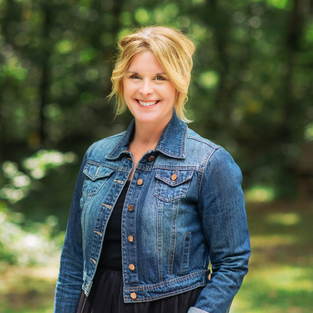 Alison Broderick smiles in a denim jacket against a green, leafy background.