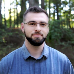 Man with beard and glasses in forest setting, wearing blue shirt.